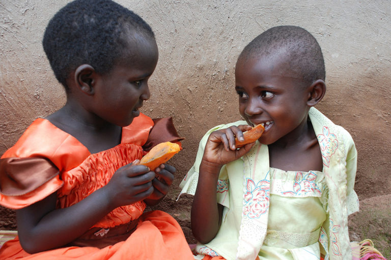 A little girl wearing an OFSP colored dress nibbles on sweetpotato with her friend. Children under five are particularly susceptible to vitamin A deficiency. Photo by Helen Keller International